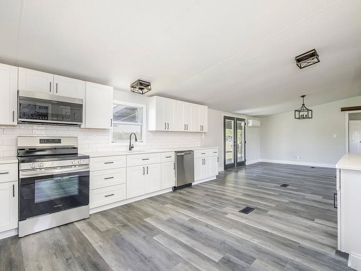 Modern kitchen with sleek white cabinets, stainless steel appliances, and light gray hardwood floors. An open layout leads to a bright dining area with large windows, creating an airy and inviting atmosphere.