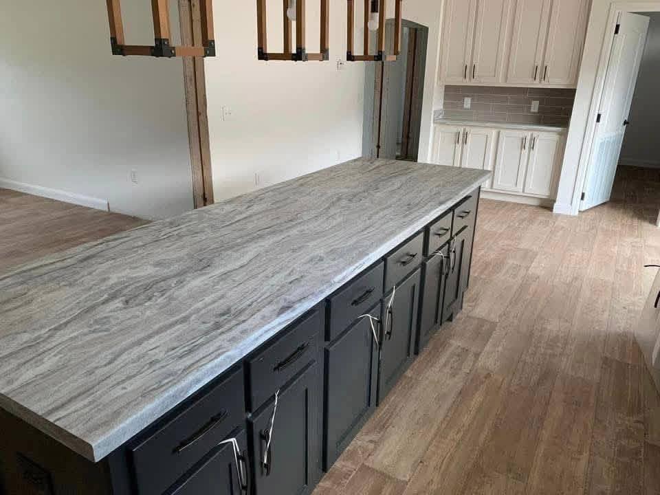 Modern kitchen featuring a long, gray marble island with dark cabinets and sleek handles, wood flooring, light hanging from ceiling, and white cabinetry.