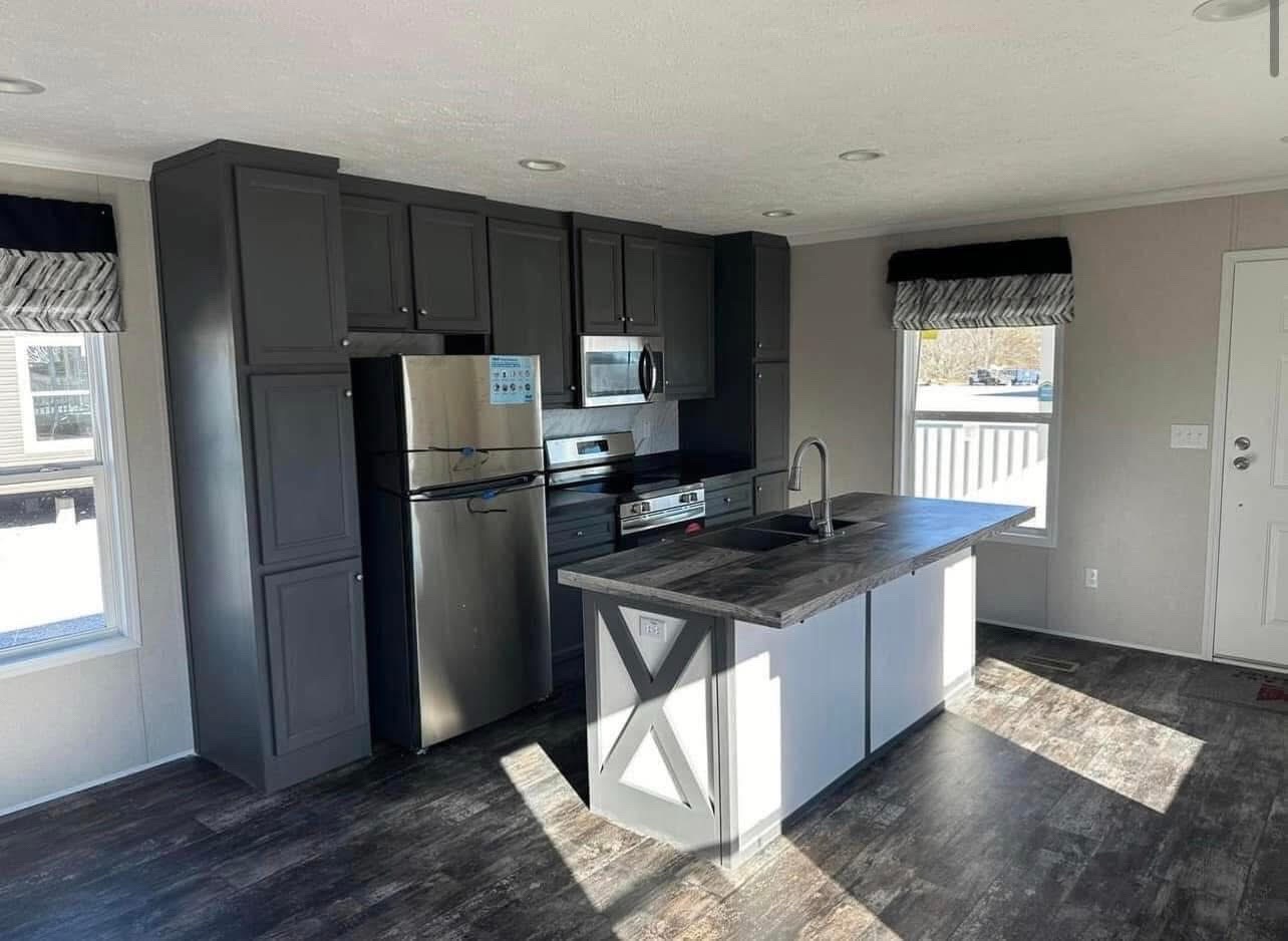 A modern kitchen with dark cabinets, stainless steel appliances, and a central island with a sink. Bright natural light streams through large windows.