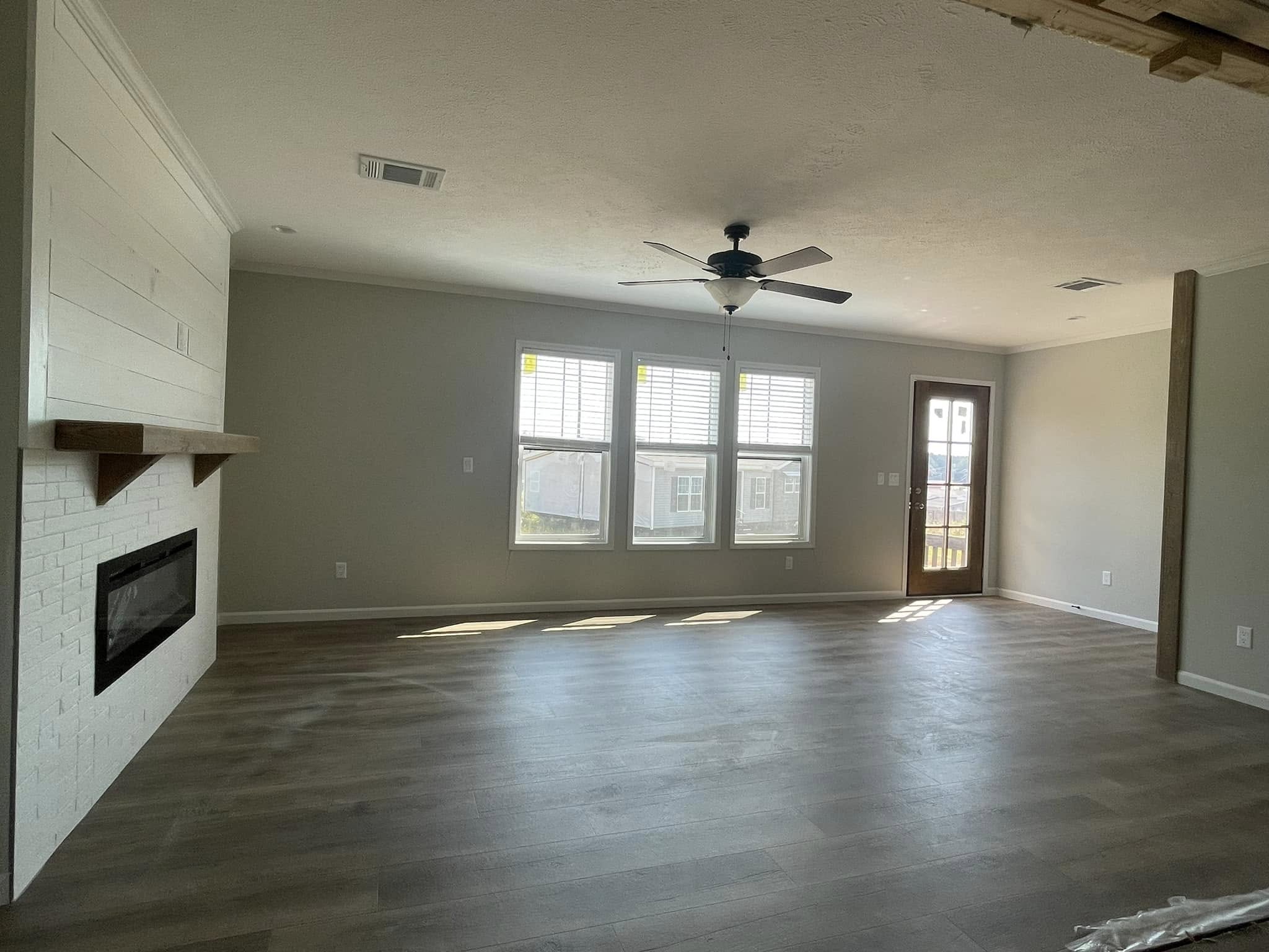 Bright, empty living room with light wooden floors, a ceiling fan, three large windows, a door to the right, and a white brick fireplace on the left.