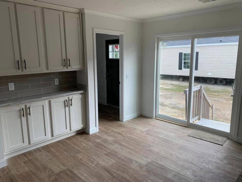 Bright room with wooden floor, white cabinets, and sliding glass doors opening to a porch. Outside view shows a parked mobile home. Calm, minimalist vibe.