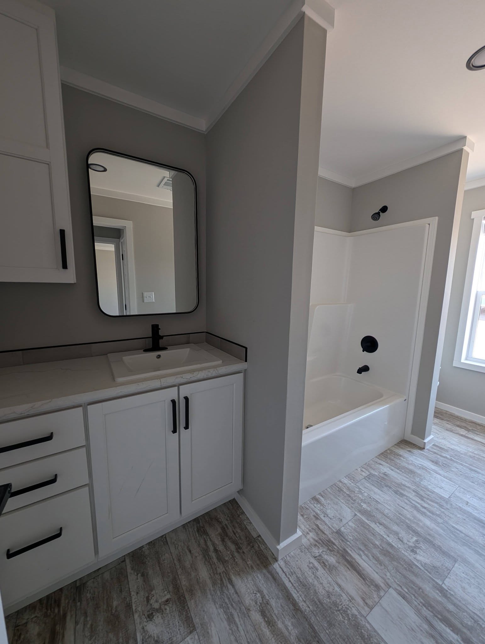 Modern bathroom with gray walls features a rectangular mirror above a white vanity with black fixtures. A bathtub and shower are beside it, exuding a sleek, minimalist vibe.