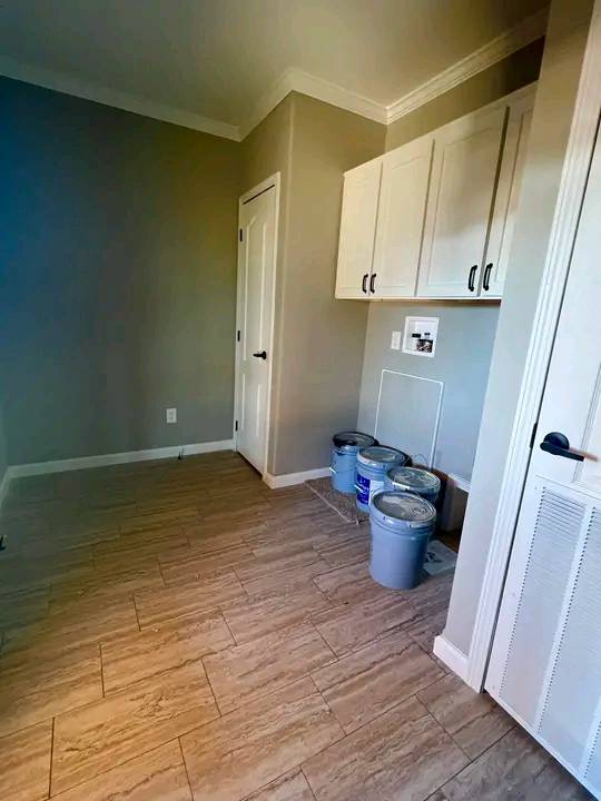 Laundry room with beige walls, tile flooring, and white cabinets. Three paint cans are on the floor beneath the cabinets, suggesting a renovation in progress.