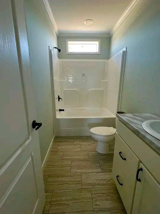 Narrow bathroom with light-colored walls, a tub-shower combo, and a small window above. A toilet and a sink with a cabinet are on the right. Calm, simple vibe.