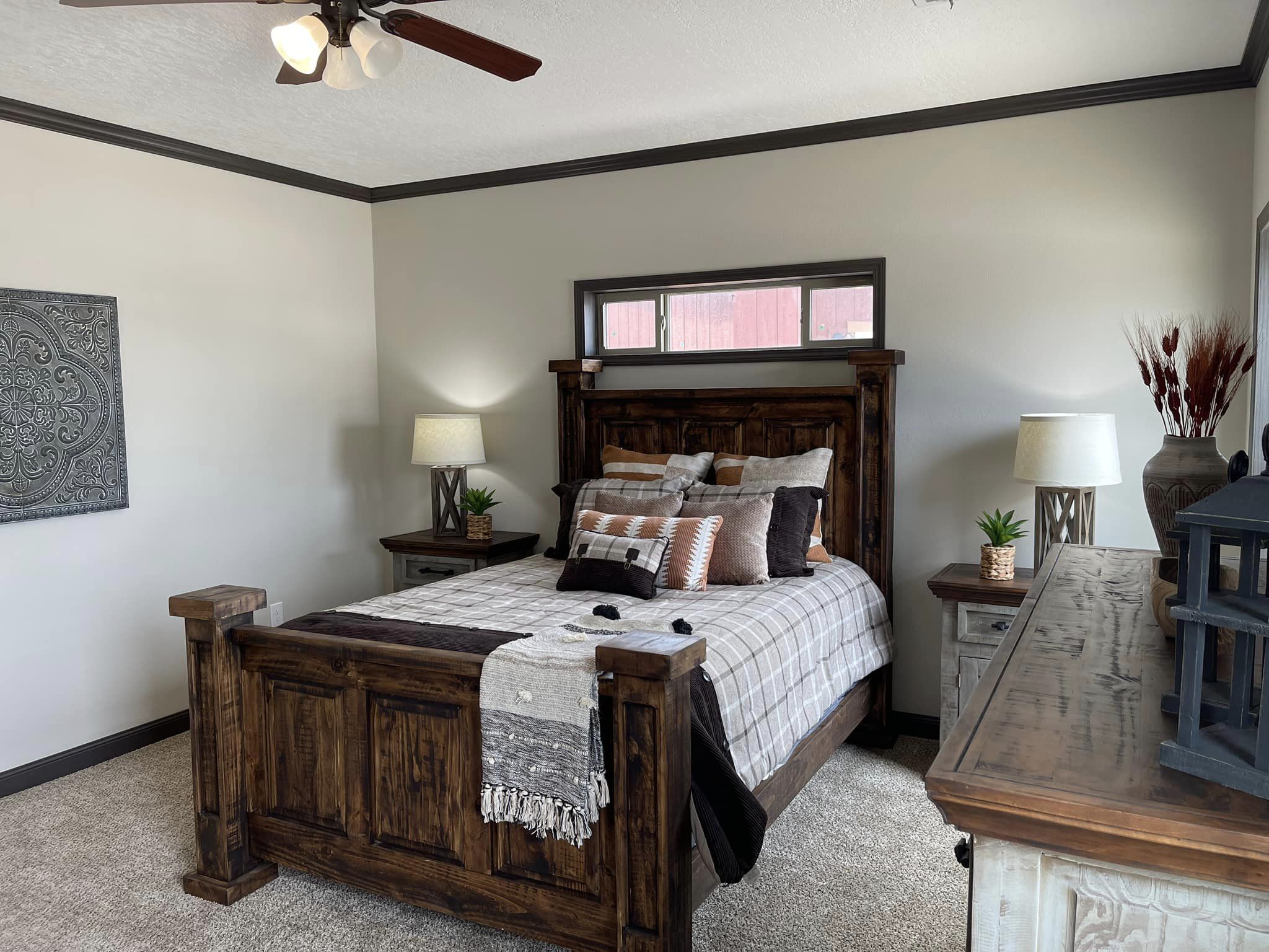 Cozy bedroom with a rustic wooden bed, plaid bedding, and assorted pillows. Nightstands with lamps and plants sit on each side. Neutral tones, calm ambiance.