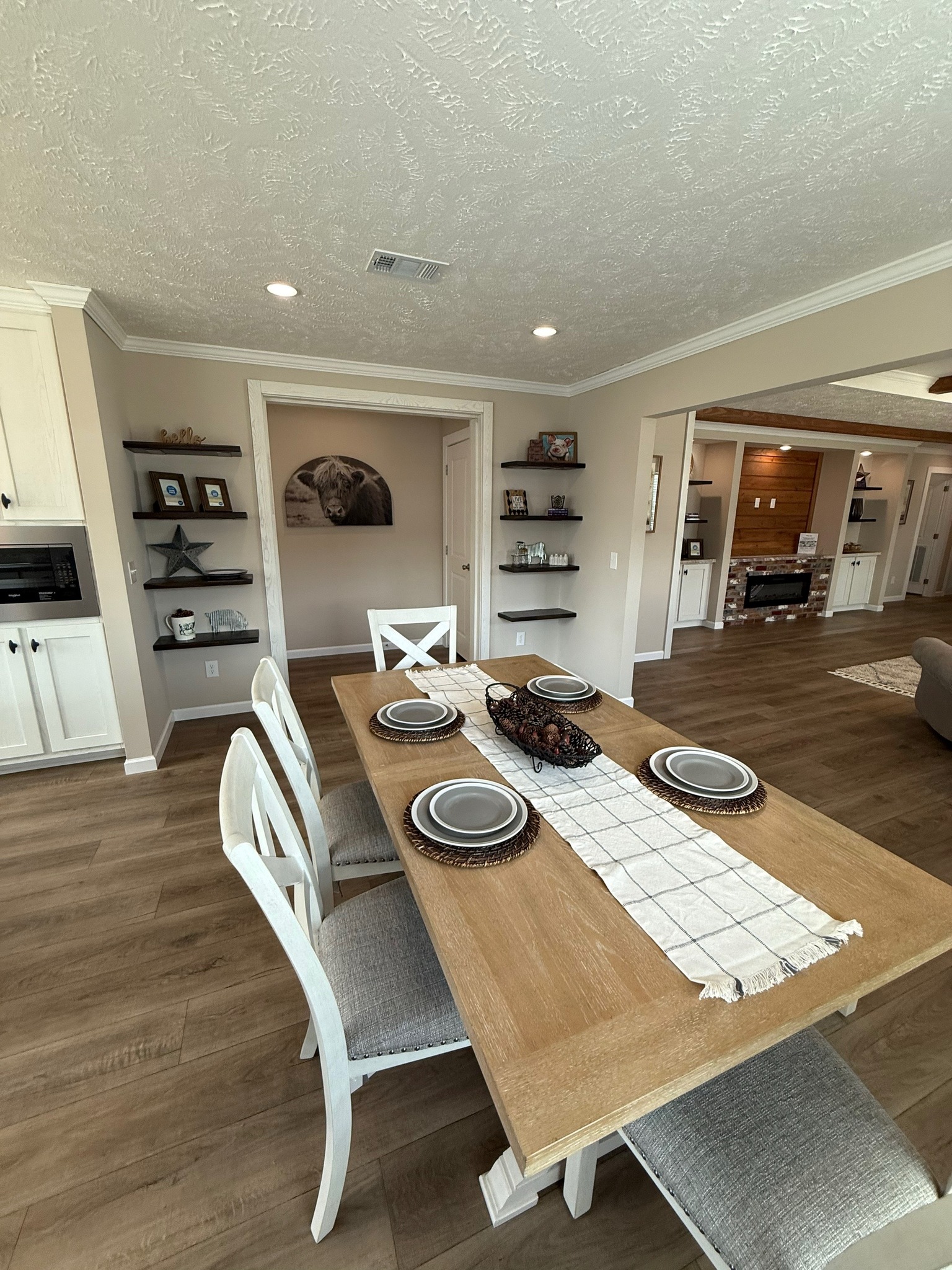 Warm dining room with a wooden table set for six; plates, woven decor, white chairs. Background: cozy living area, fireplace, animal artwork.