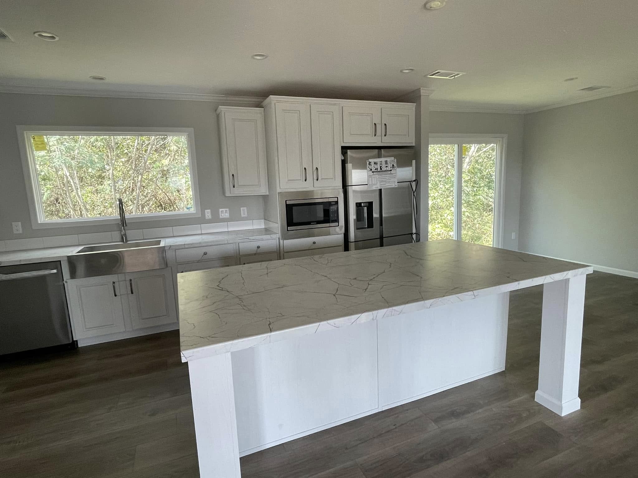 Modern kitchen with white cabinets, stainless steel appliances, and large marble island. Sunlit through a window and glass doors, creating a serene ambiance.