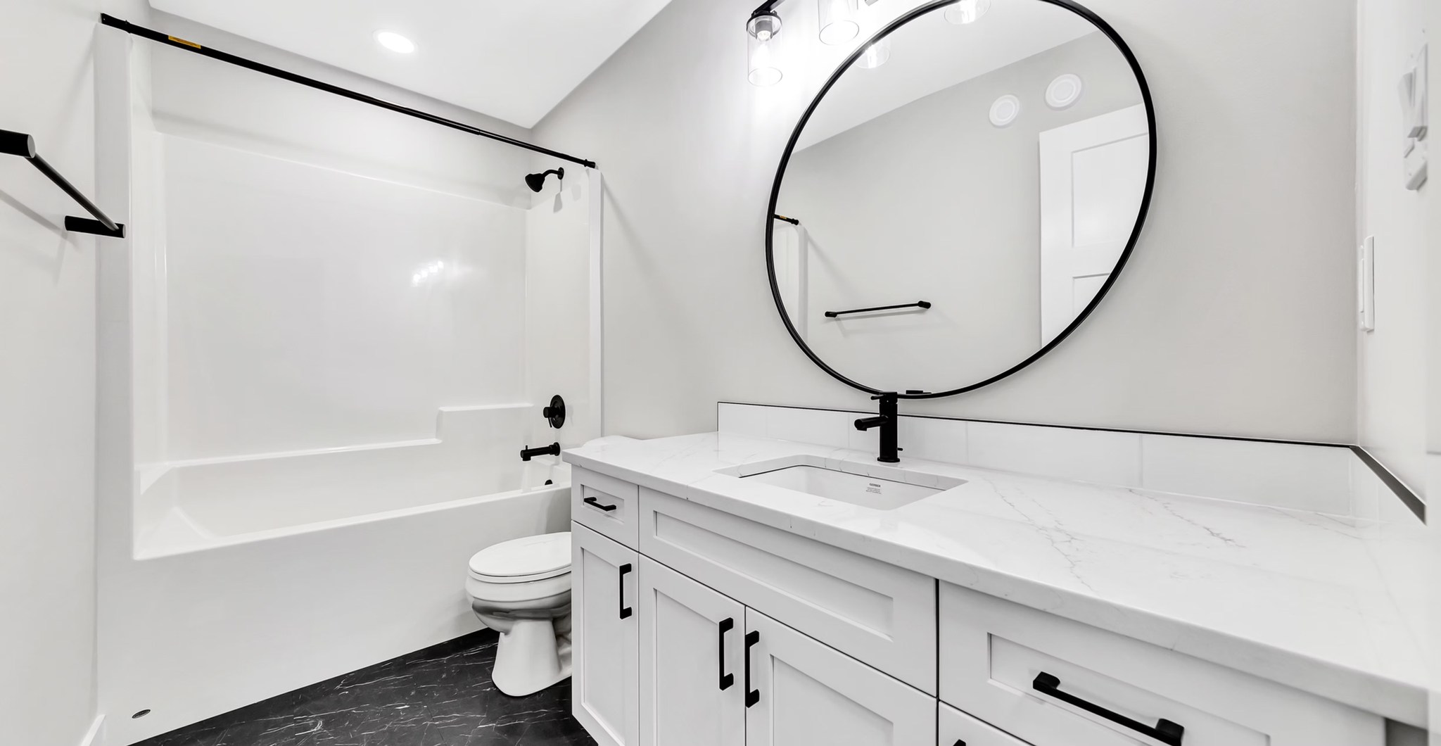 Modern bathroom with a white bathtub and toilet on the left. A large round mirror hangs above a white vanity with black fixtures, exuding a sleek, clean ambiance.