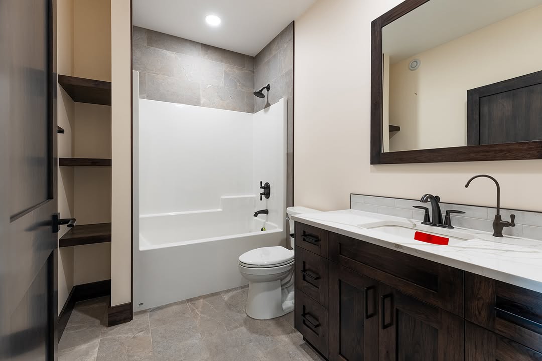 Modern bathroom with a dark wood vanity, white countertop, and matching framed mirror. Features a white bathtub with overhead shower, and beige walls.