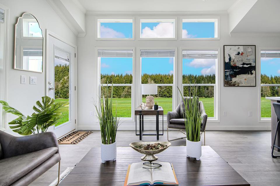 A bright living room with large windows showcasing a scenic view of a green field and blue sky. The room has modern furniture, plants, and abstract art.