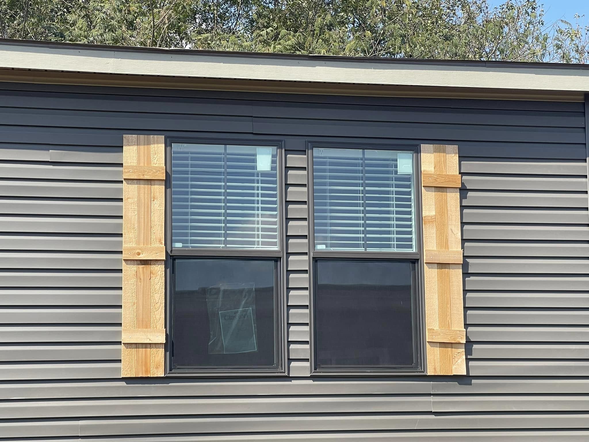 A double window with white blinds is set in dark gray siding, featuring rustic wooden shutters. Trees are partially visible above, under a blue sky.