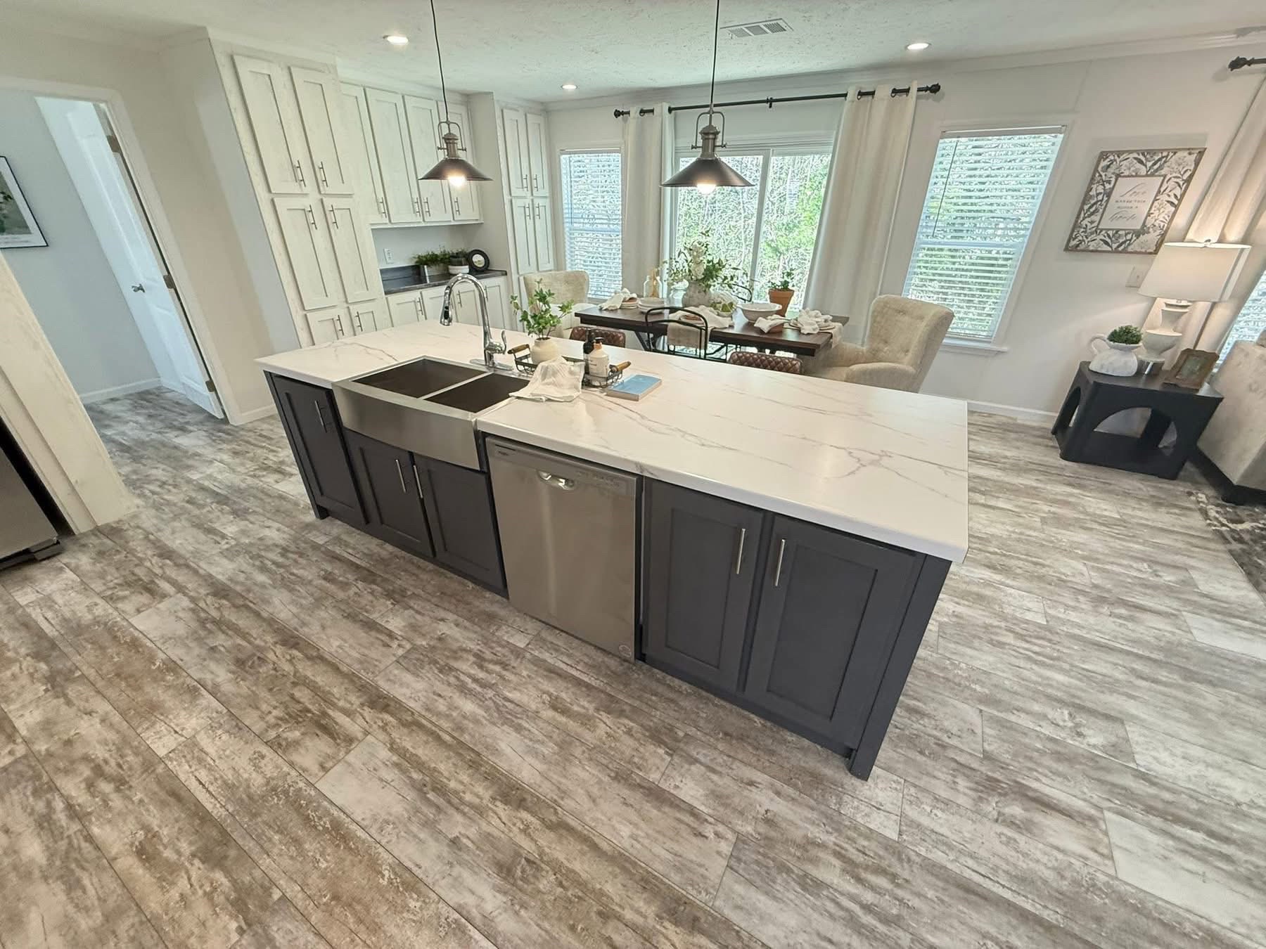 Bright kitchen with gray cabinets and marble island, stainless steel sink, and hanging pendant lights. Cozy dining area with large windows in the background.