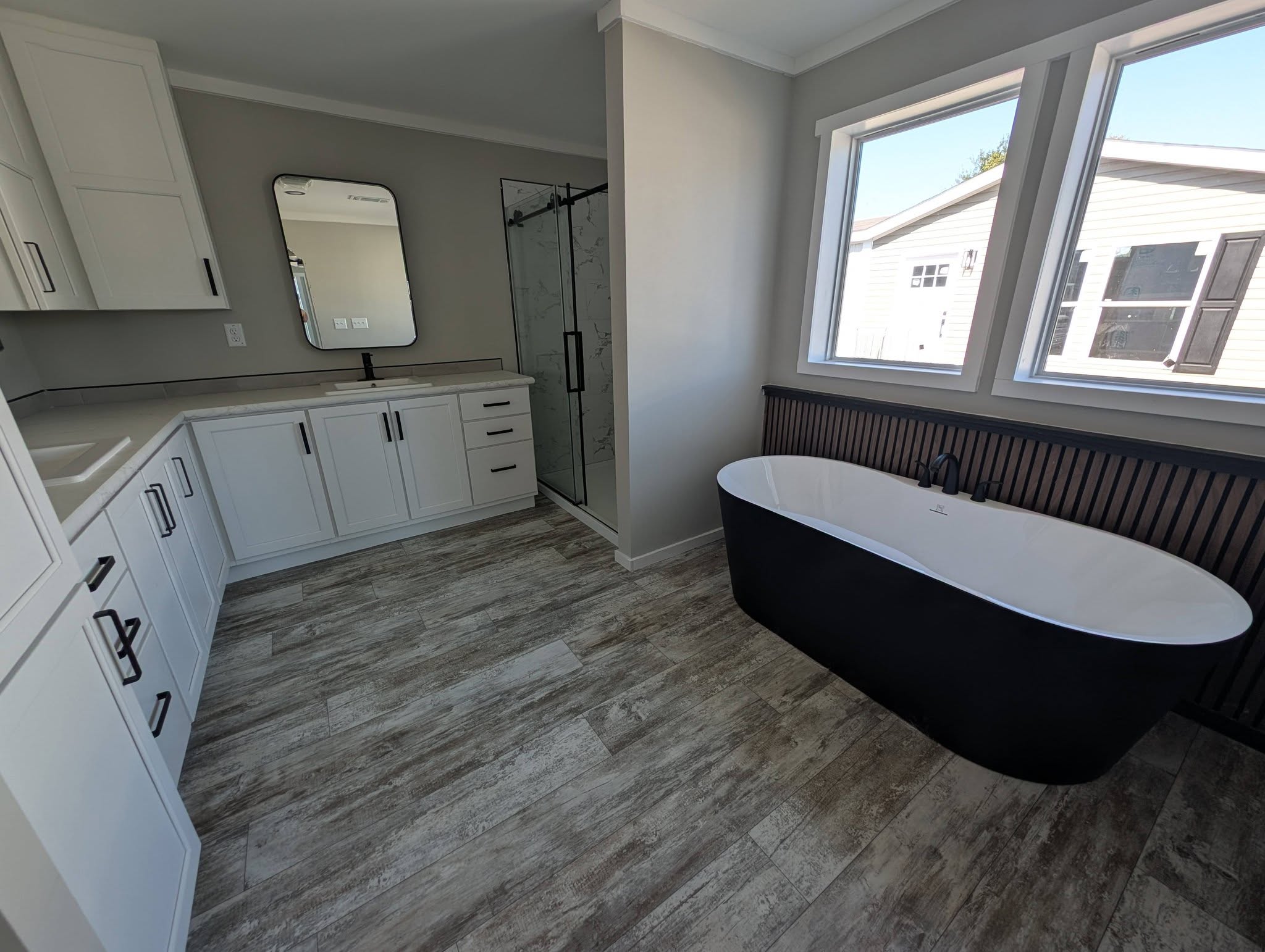 Modern bathroom with light wood flooring, featuring a freestanding black and white tub, large windows, white cabinets, and a walk-in glass shower.