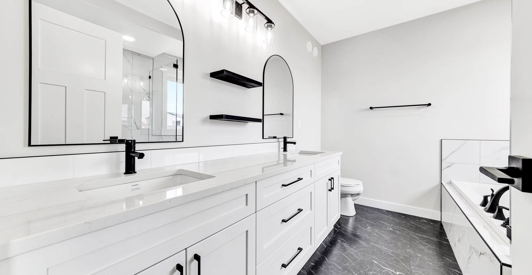 Modern bathroom with dual white sinks and black fixtures on a large vanity. Arched mirrors, black shelves, and a bathtub on dark tile flooring.
