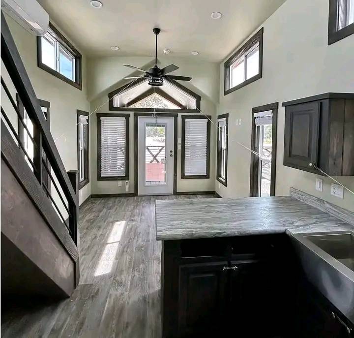 Modern, sunlit tiny house interior with high ceilings and loft. A fan hangs from the ceiling, surrounded by windows. Dark wood cabinets contrast with light walls.