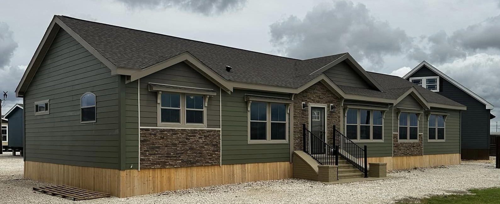A modern, single-story modular home with dark green siding and stone accents under a cloudy sky. The house features multiple windows and a small porch.