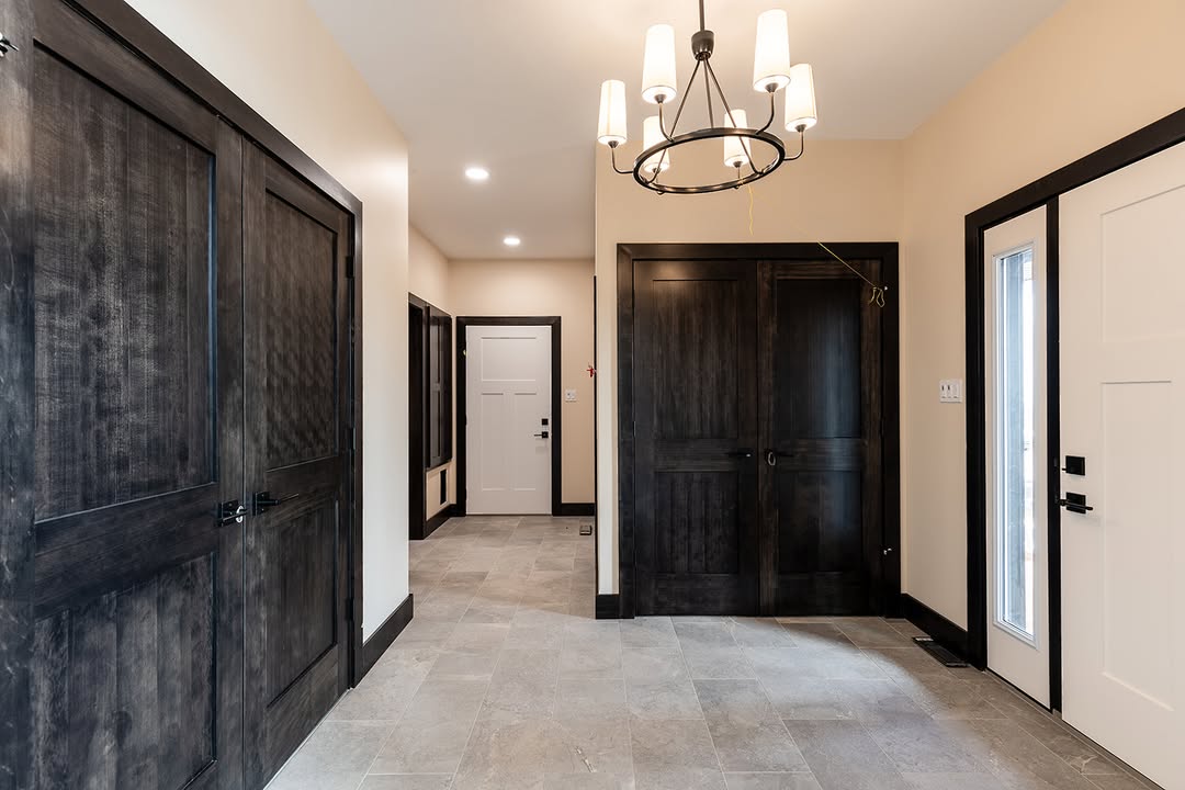 A modern hallway with beige walls, dark wooden doors, and light gray tiled floor. A black chandelier with white lampshades hangs from the ceiling, adding elegance.