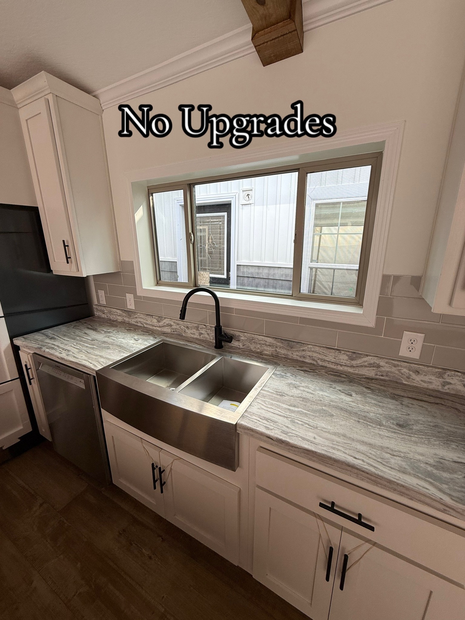 Modern kitchen with white cabinets, gray marble countertop, and a large stainless steel sink beneath a window. Text reads "No Upgrades." Bright, clean ambiance.