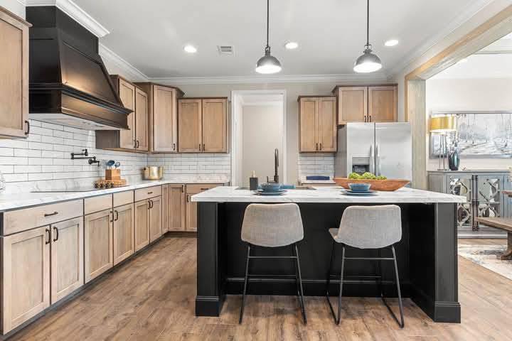 Modern kitchen with light wood cabinets, white subway tile backsplash, and dark kitchen island. Two gray chairs, pendant lights, and stainless steel appliances create a sleek, inviting atmosphere.