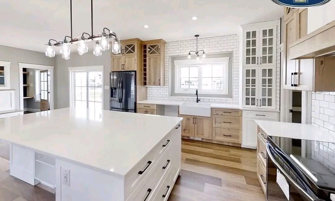 Modern kitchen with a spacious white island, pendant lights, farmhouse sink, subway tile backsplash, natural wood cabinets, black fridge, and large window.