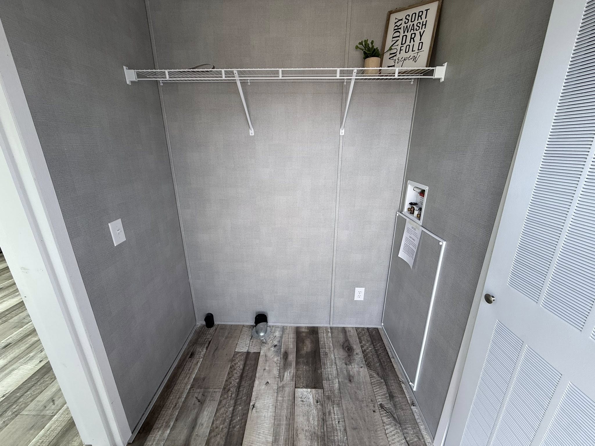 Empty laundry room with gray walls and wood-patterned floor. A wire shelf holds a potted plant and a sign reading "Sort, Wash, Dry, Fold, Repeat."