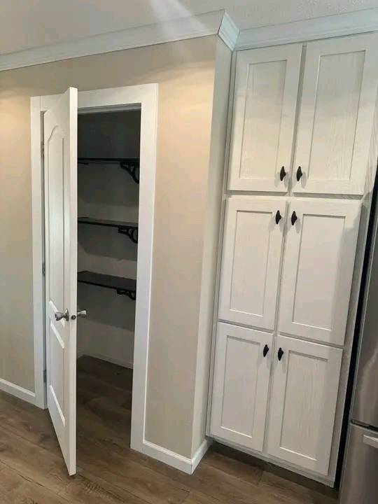 Open pantry door reveals dark shelves against cream walls, adjacent to white cabinetry with black handles. Hardwood floor adds warmth to the kitchen.