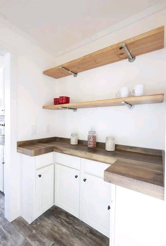 Simple corner kitchen with white walls and rustic wood elements. Two wooden shelves hold red bowls and white mugs; jars are on the wooden counter.