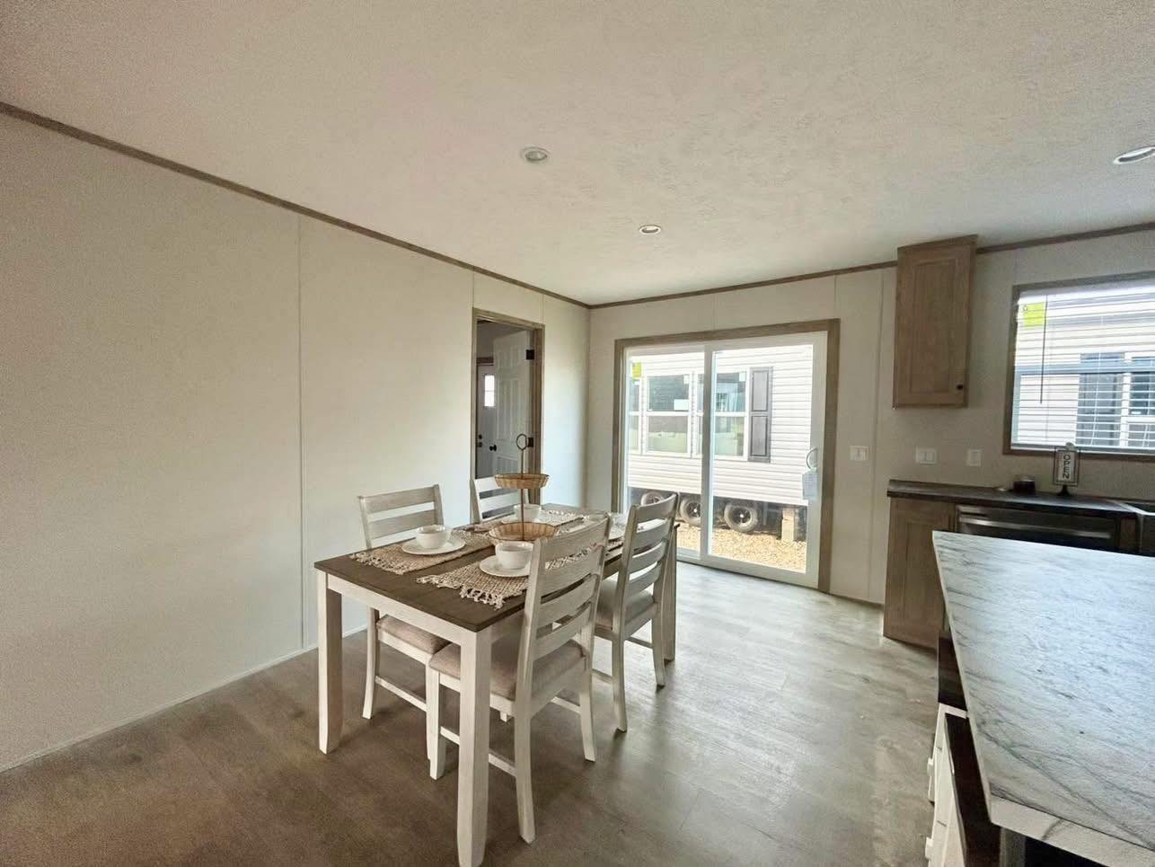 Bright dining area with a rustic wooden table set for four, next to large sliding glass doors. The space feels open and inviting with light walls.