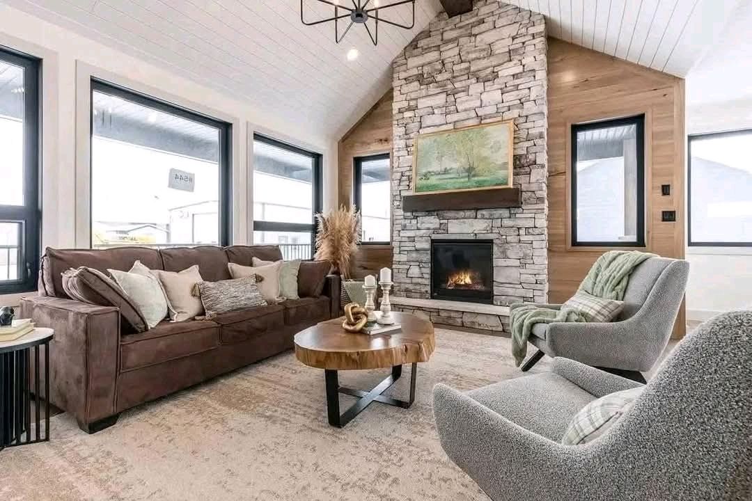 Cozy living room with a stone fireplace, brown sofa, and modern chairs. Large windows provide natural light. A wooden coffee table adds warmth.