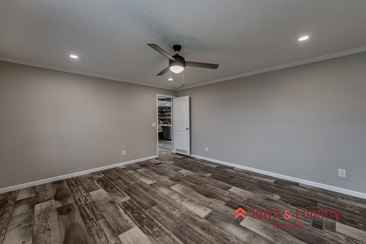 Empty room with wood flooring, beige walls, and a ceiling fan. An open door reveals a hallway and cabinets. Recessed lighting creates a warm ambiance.