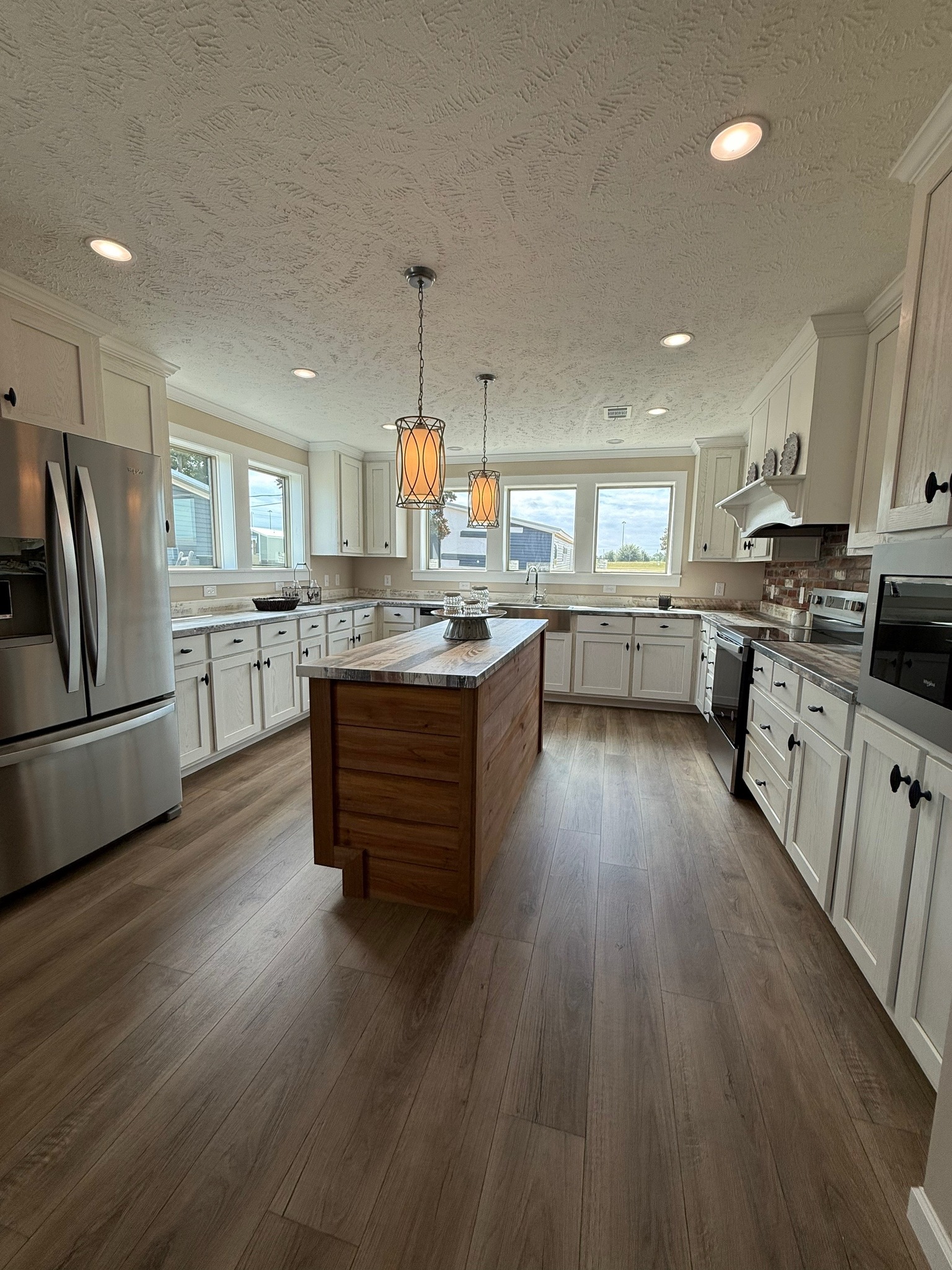 Spacious kitchen with wooden floors, center island, and stainless steel appliances. Pendant lights hang above the island. Large windows offer natural light.