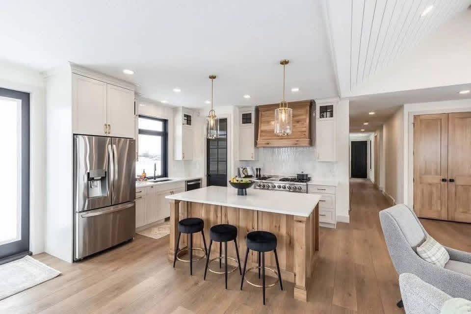 Modern kitchen with a bright, airy feel. Features a large island with black stools, stainless steel appliances, and pendant lights. Warm wood tones and sleek finishes.