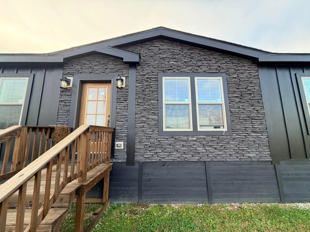 Front view of a modern house with dark stone facade, wooden door, stairs, and two large windows with white blinds. The sky is overcast, adding a cozy feel.