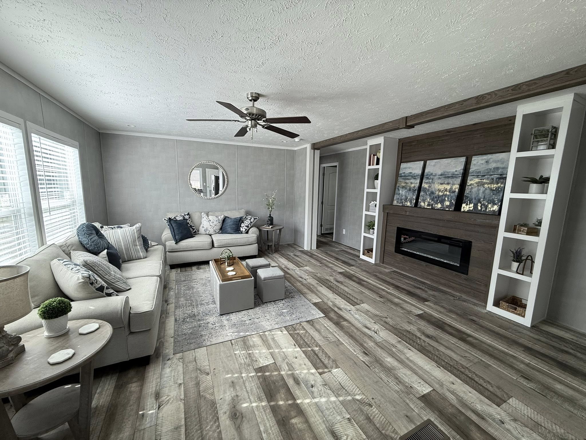 Modern living room with a neutral palette, featuring a gray sectional sofa, decorative pillows, wooden coffee table, large windows, and a wall-mounted TV.
