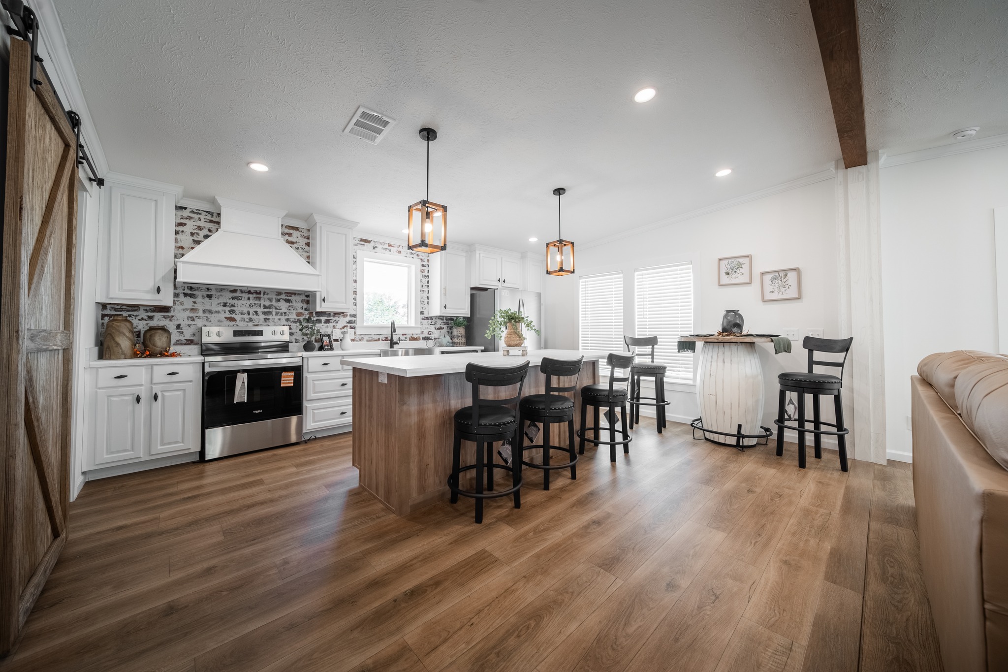 Spacious kitchen with warm wood flooring, white cabinetry, brick backsplash, and a central island with five black stools. Cozy and modern atmosphere.