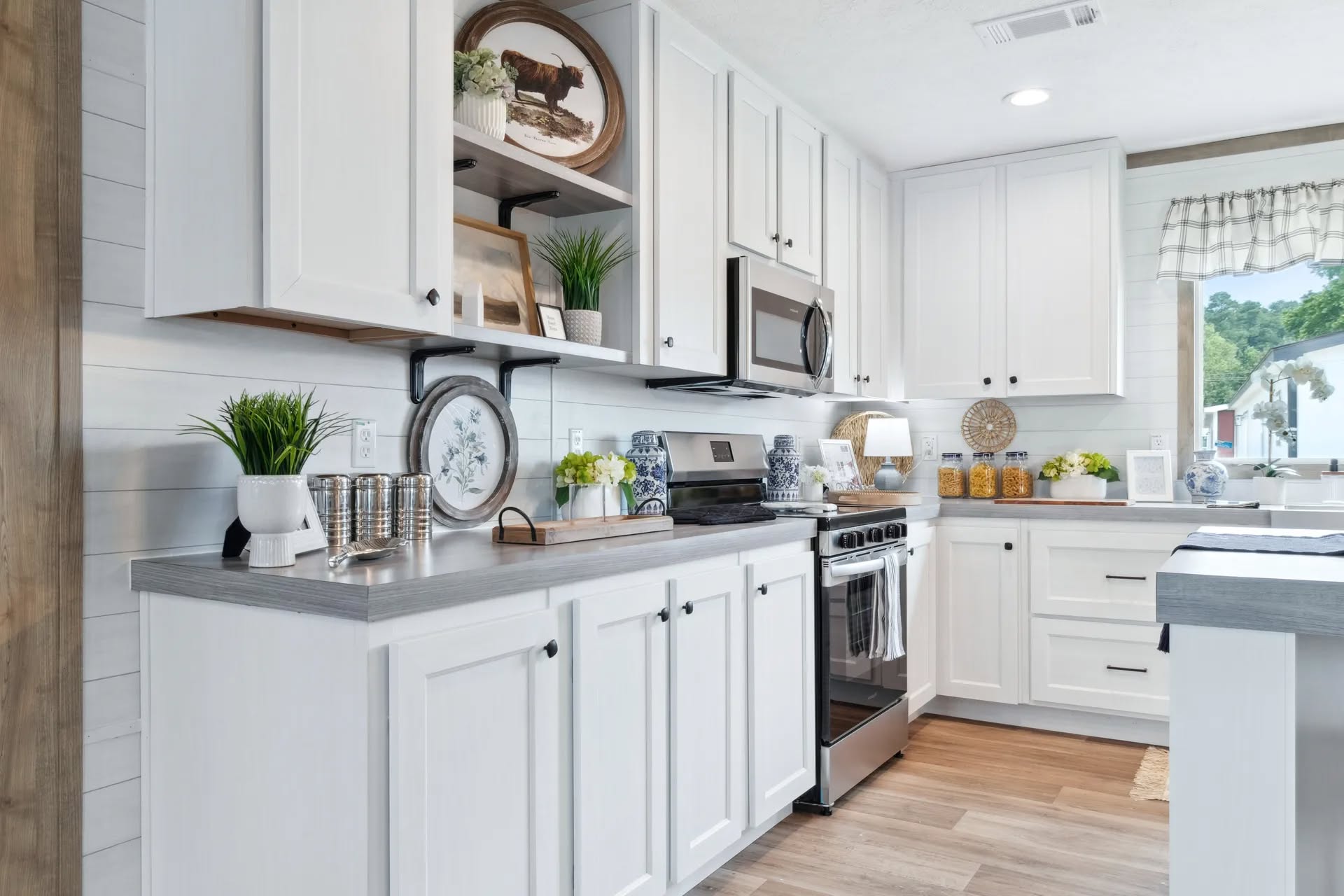 Bright, modern kitchen with white cabinets, stainless steel appliances, and rustic decor. Open shelves display plants and ceramics, creating a cozy, inviting feel.