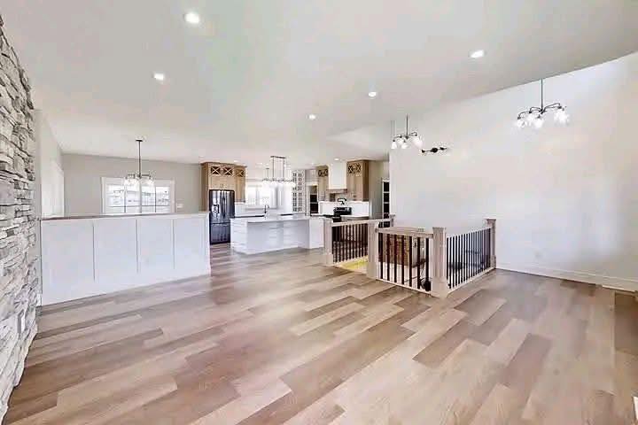 Spacious modern living area with light wood flooring, white walls, and ceiling lights. Open kitchen in background, featuring a large island and elegant fixtures.