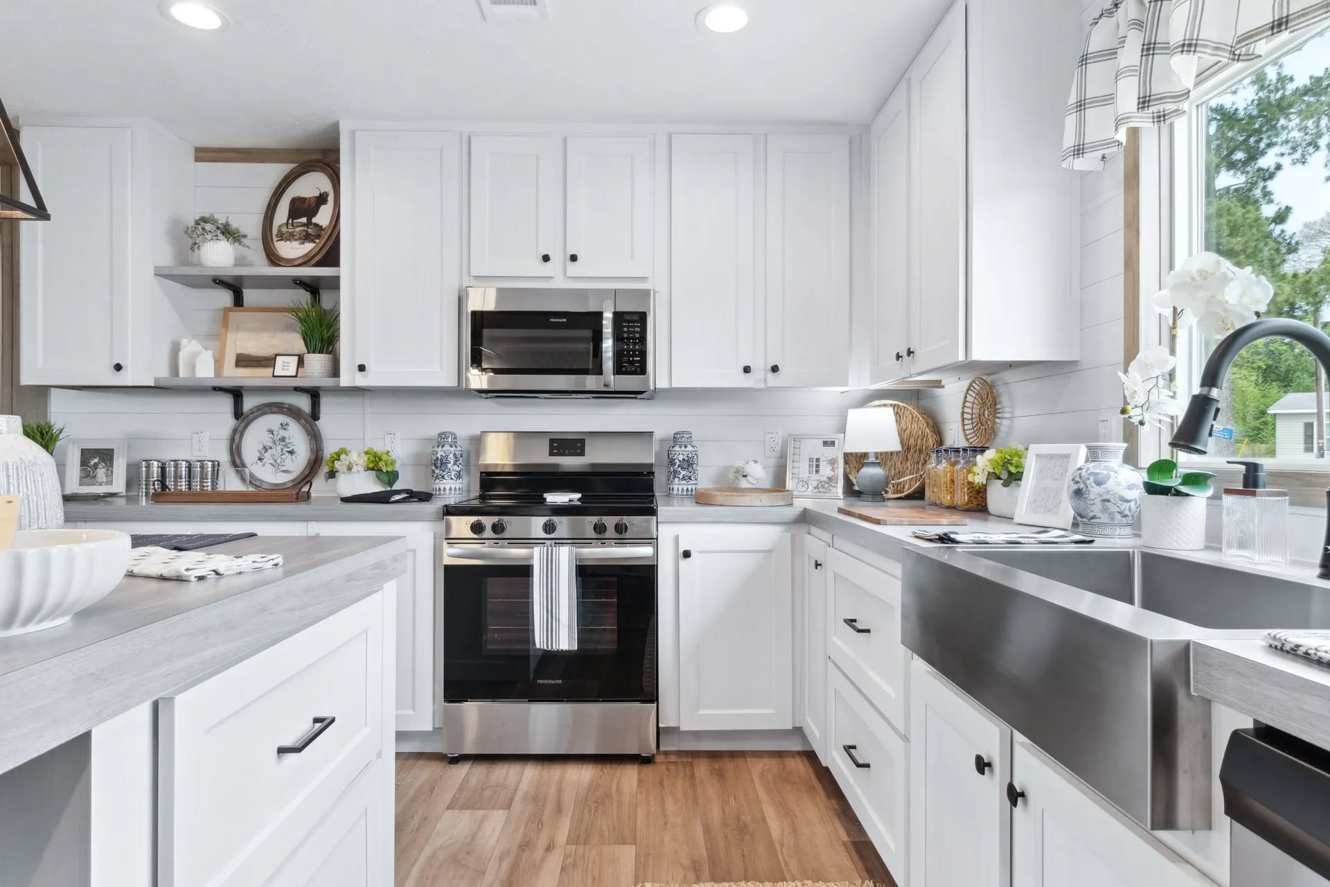 Bright modern kitchen with white cabinets, stainless steel appliances, and wooden flooring. Open shelves display decor; a large window brings in natural light.