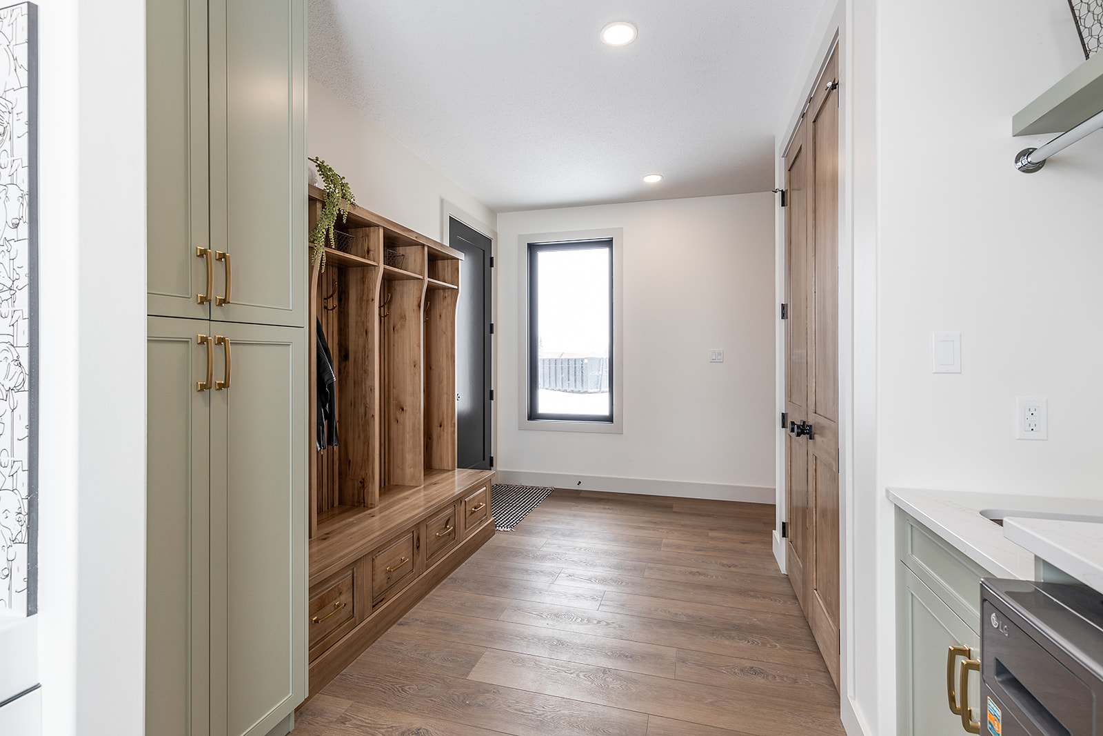 A bright mudroom with light wood floors, sage green cabinets, and a wooden bench with hooks and storage. A window and wooden doors add warmth.
