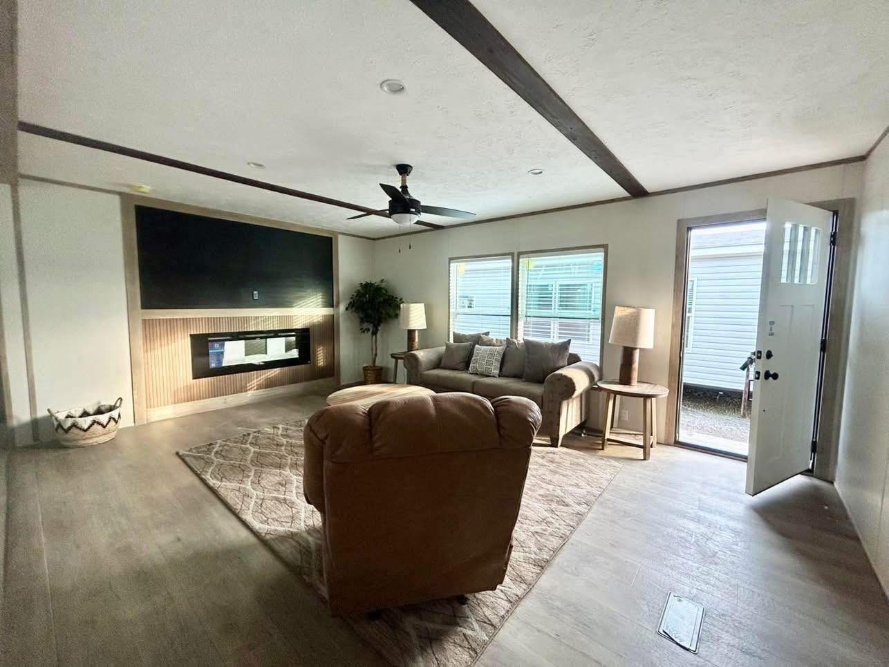 Cozy living room with a tan couch, armchair, and a fireplace. A large window and open door let in natural light. Neutral tones create a warm atmosphere.