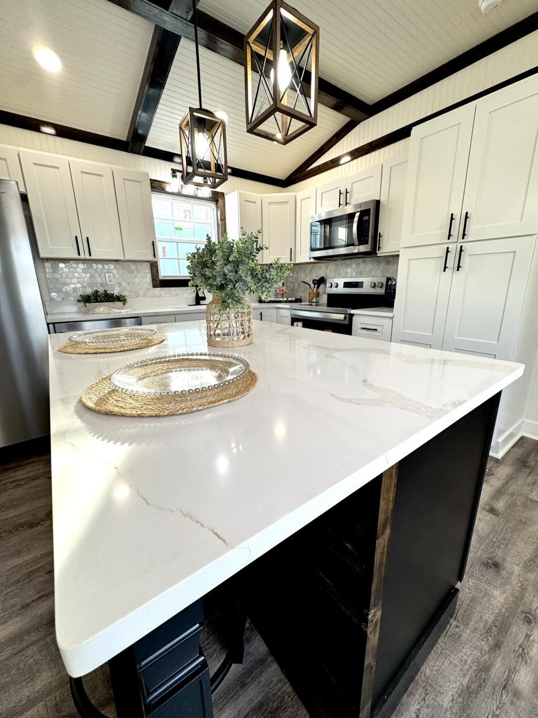 Spacious kitchen with a marble island, wicker placemats, and greenery centerpiece. White cabinets, stainless steel appliances, exposed beams, and elegant pendant lights convey a modern, inviting atmosphere.
