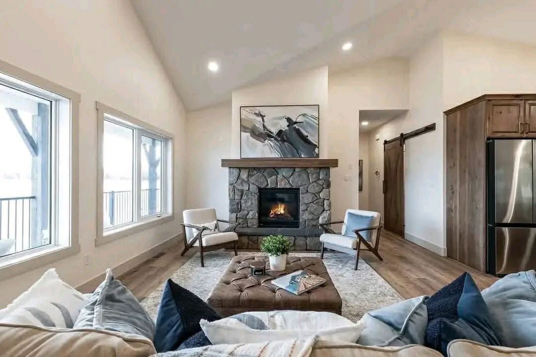 A cozy living room with a stone fireplace, abstract art, and two armchairs. A cushioned coffee table holds a plant and books. Warm and inviting.