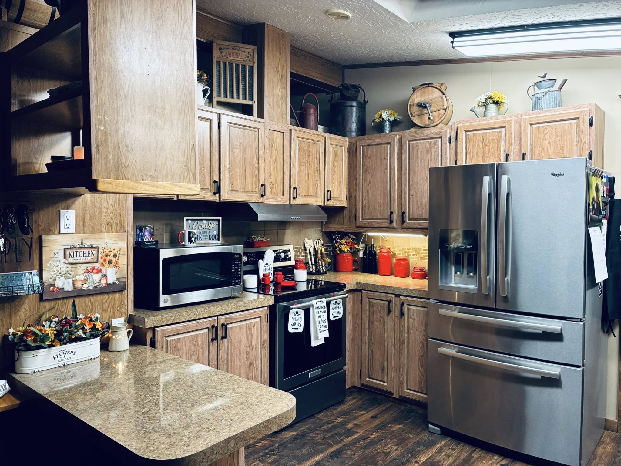 Warm, rustic kitchen with wooden cabinets, stainless steel fridge, and dark stove. Red accents, floral decor, and cozy ambiance fill the room.