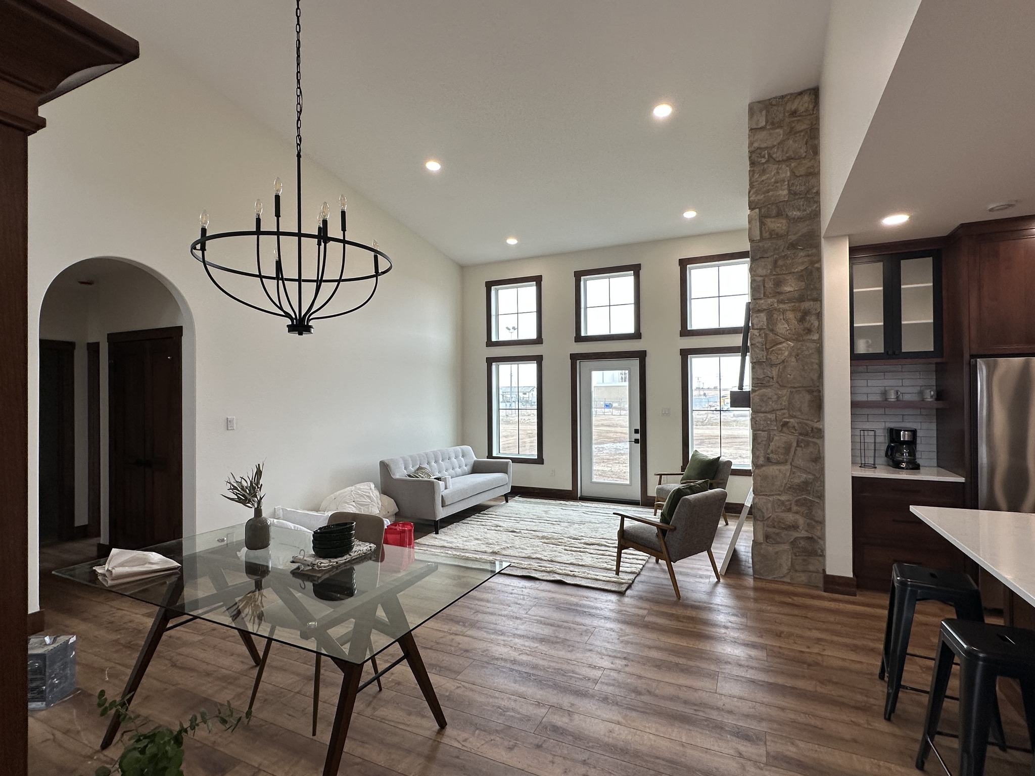 A bright living room with large windows, a gray sofa, and an armchair. The open space includes a dining table with a chandelier above and wood floors.