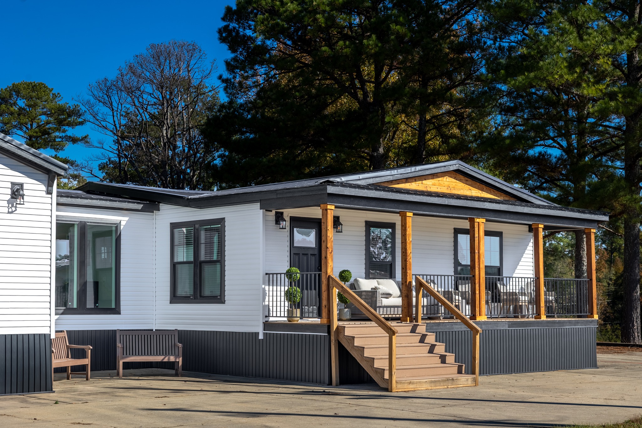 Modern white manufactured home with a covered wooden porch, surrounded by trees. The setting is calm and sunny, featuring sleek, minimal design elements.