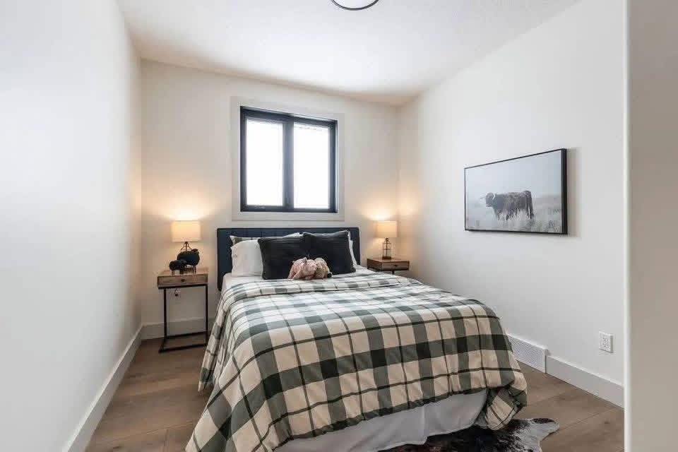Cozy bedroom with plaid bedding, soft lighting from two bedside lamps on wooden tables, and a window above the bed. A cow painting adorns the wall.