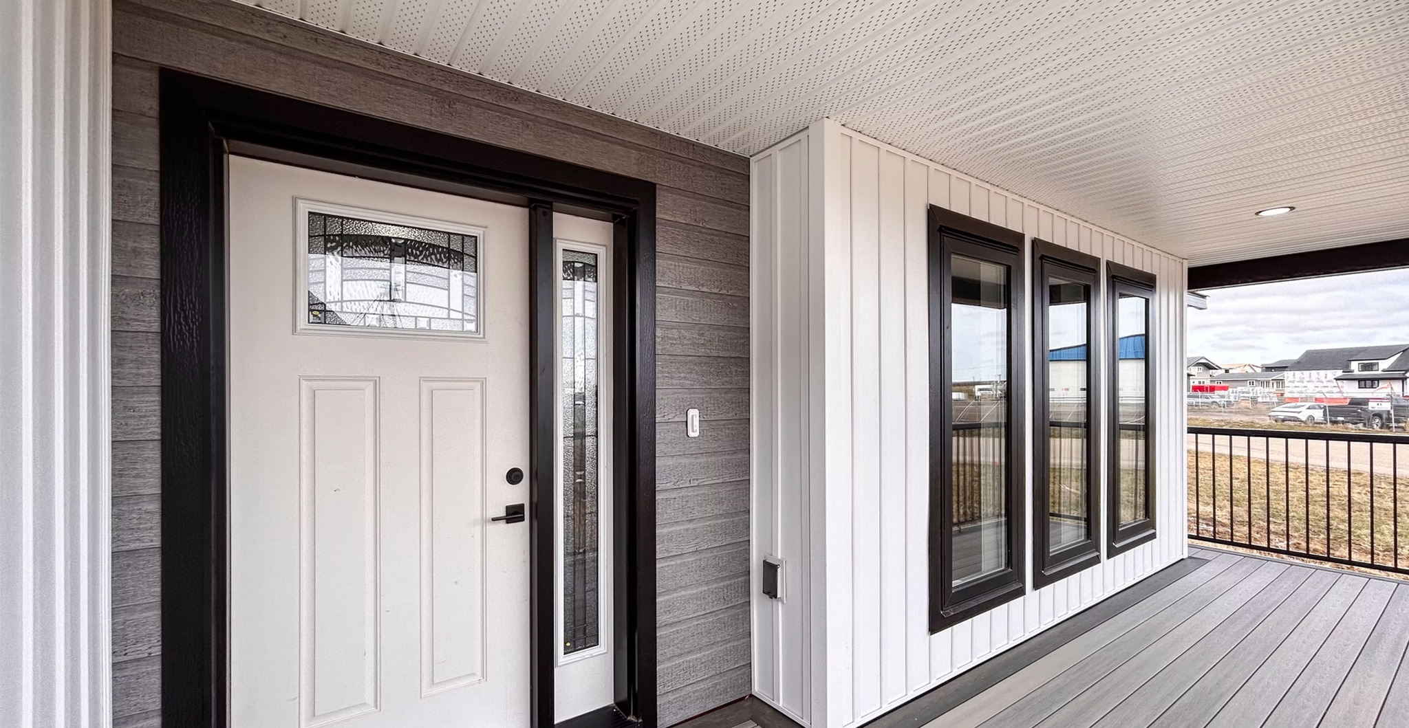 Modern house entrance with a white door featuring decorative glass panels, surrounded by gray and white siding. Multiple windows and a spacious porch create an inviting tone.