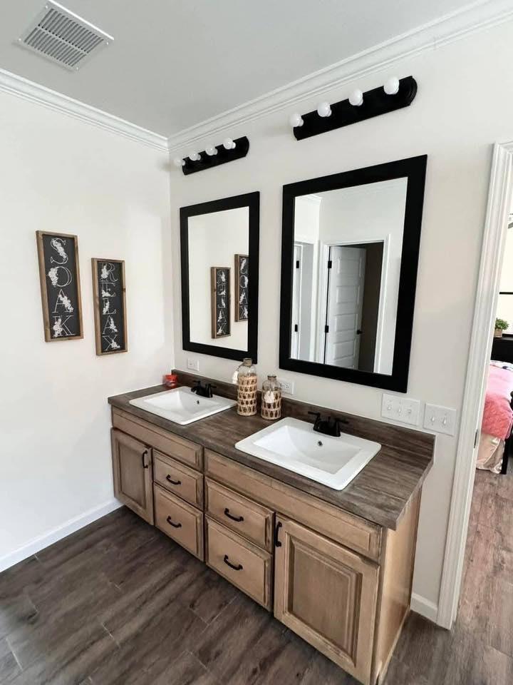 Dual-sink bathroom vanity with wooden cabinets, black-framed mirrors, and wall-mounted lights. Three decorative wall signs read “Soak,” “Relax.”