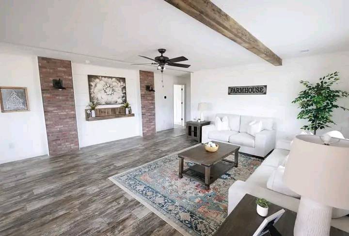 Bright living room with a farmhouse theme, featuring a white sofa, rustic wood beams, brick accents, wooden table, ceiling fan, and a colorful rug.