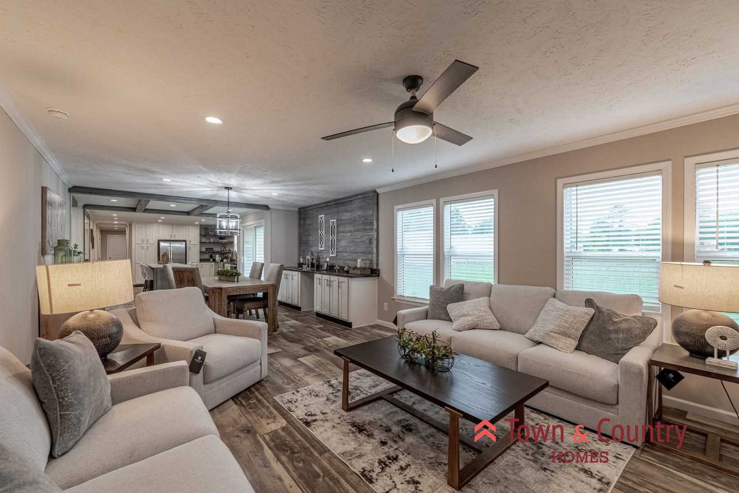 Spacious living room with modern decor, featuring comfy beige sofas, a wooden coffee table, and large windows. Open layout connects to kitchen. Cozy and inviting.